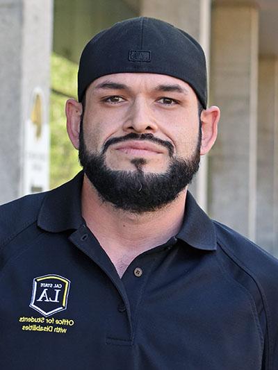 A person with a beard wearing a baseball cap backwards.