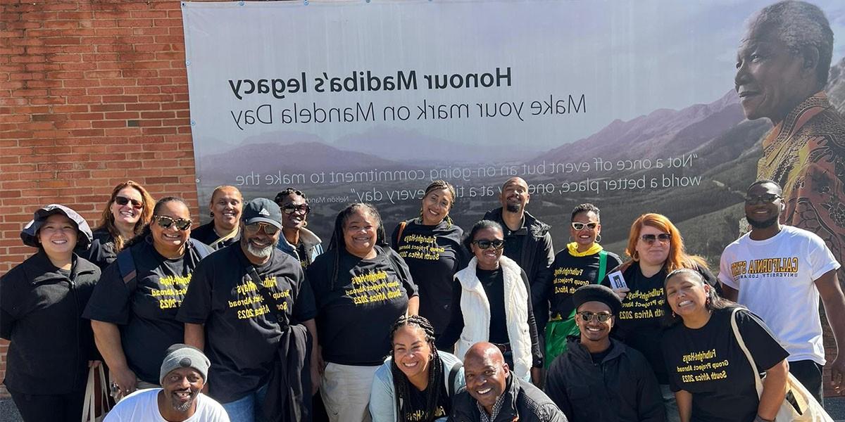 Group of students and faculty in Africa in front of poster that re广告 Honour Madiba's legacy, make your mark on Mandela Day as part of theFulbright Hays Group Project