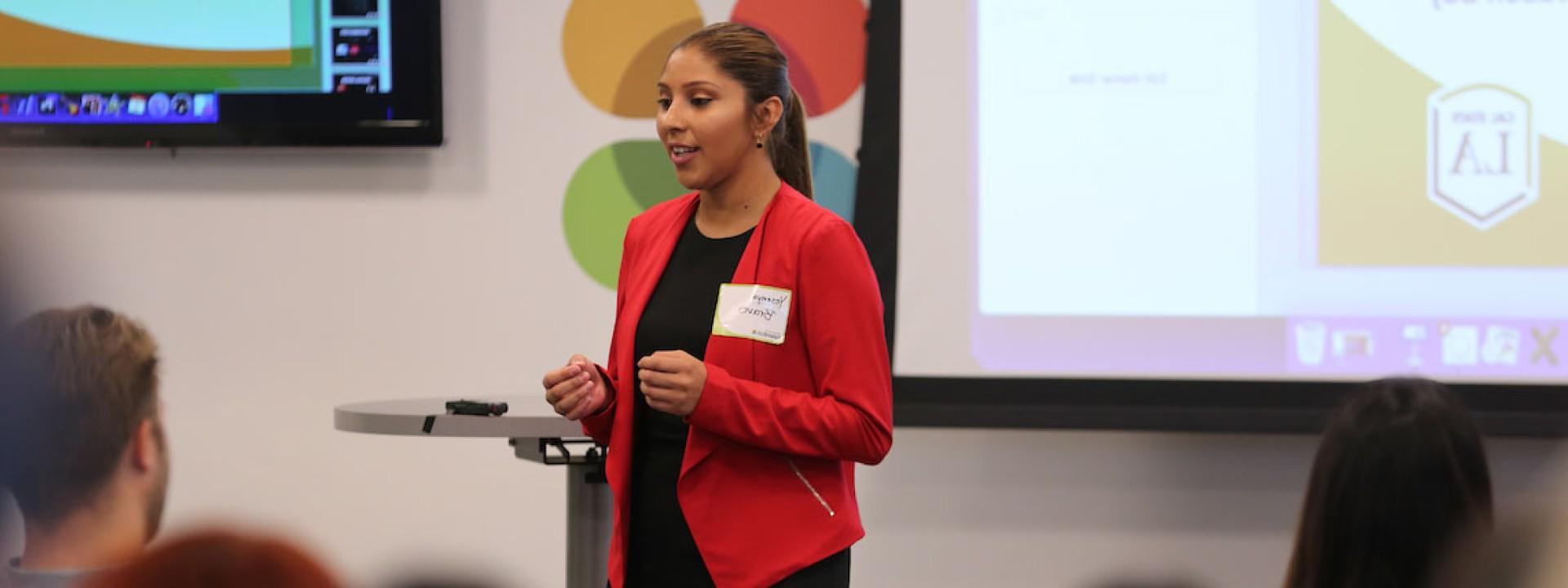 Image of a female student in a red blazer giving a presentation to her classmates. 