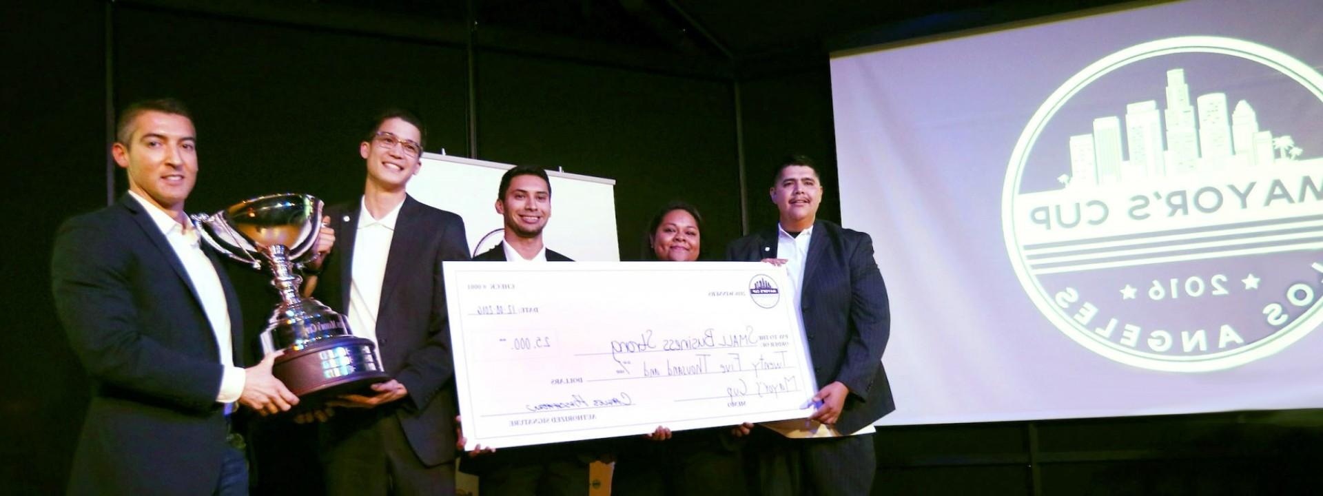 Group of Cal State LA students in business attire on a stage holding a large check near a screen that says "2016 Mayor's Cup"